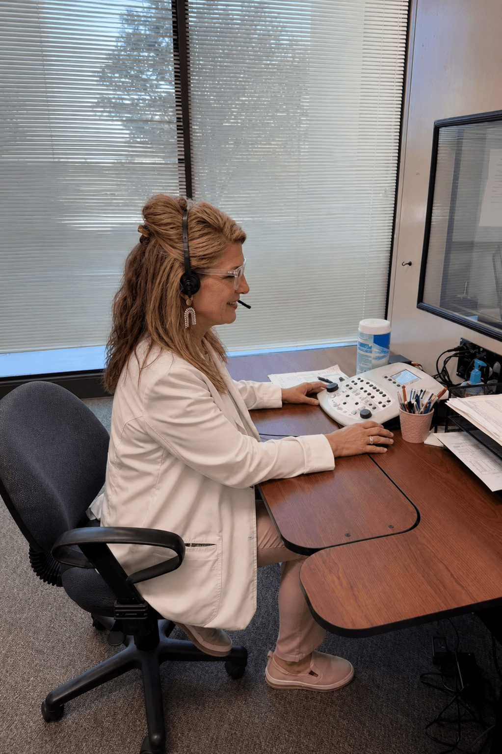 A woman in a white lab coat and headset operates an audiometer at a desk.