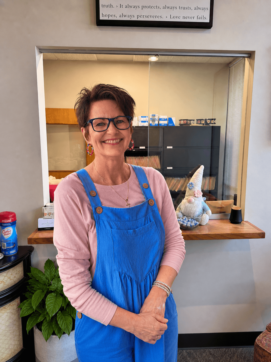 Smiling woman with short hair and glasses wearing blue overalls and a pink long-sleeved shirt.